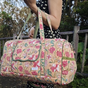 May include: A quilted duffel bag featuring a peach-colored base with a repeating floral pattern in shades of pink, white, and green. The bag includes olive green piping, two top handles, a front pocket with a flap, and a small white rectangular label.