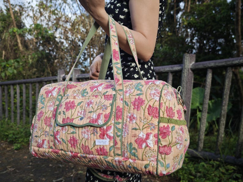 May include: A quilted duffel bag featuring a peach-colored base with a repeating floral pattern in shades of pink, white, and green. The bag includes olive green piping, two top handles, a front pocket with a flap, and a small white rectangular label.