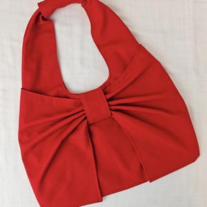 May include: A vibrant red handbag with a unique bow-like design. The bag features a single handle and a large, central bow detail. The fabric appears to be a smooth, solid color, creating a bold and eye-catching accessory.