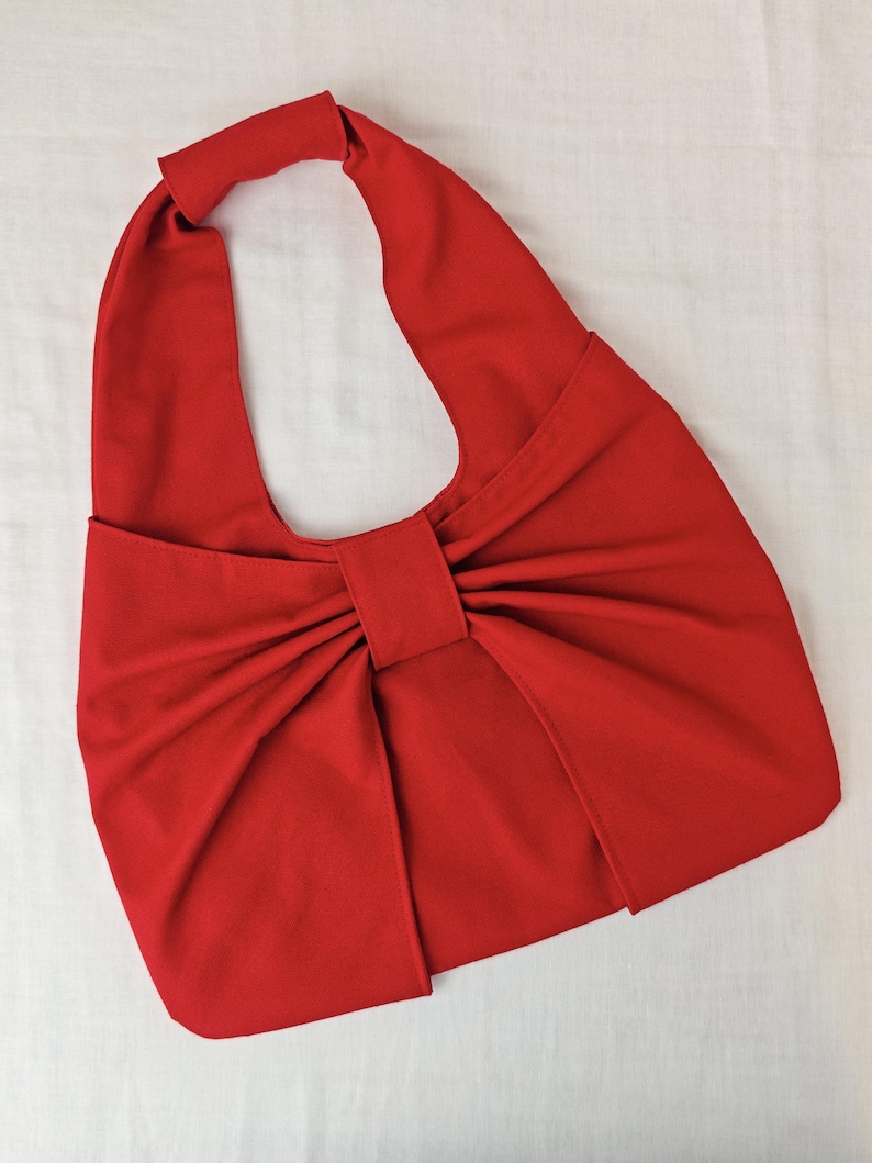 May include: A vibrant red handbag with a unique bow-like design. The bag features a single handle and a large, central bow detail. The fabric appears to be a smooth, solid color, creating a bold and eye-catching accessory.