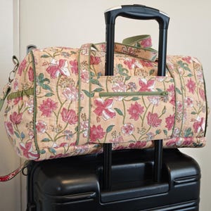 May include: A quilted duffel bag with a light peach background and a floral pattern in shades of pink, red, and green. The bag features a green zippered pocket on the front, green trim, and a strap designed to slide over a black luggage handle.