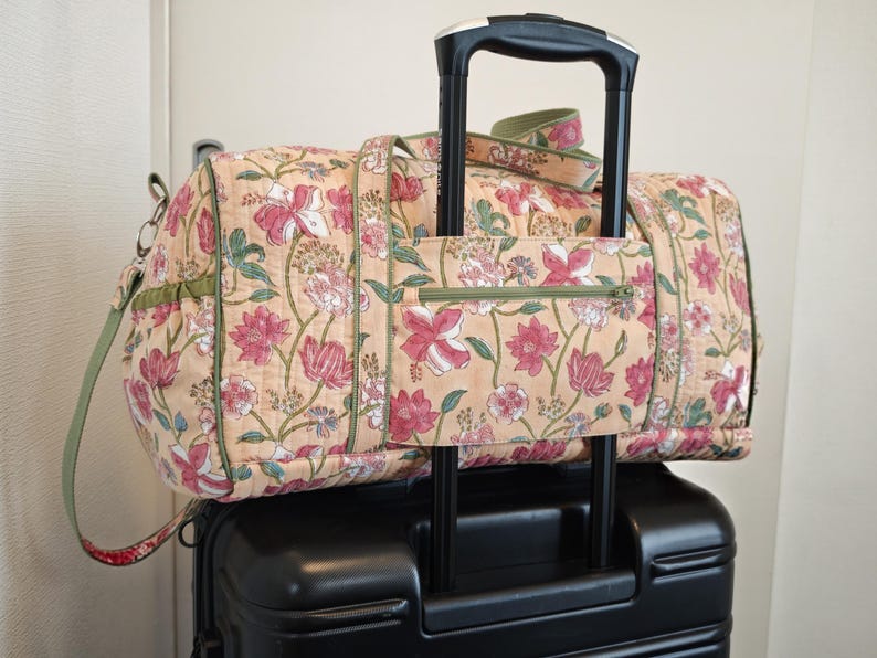 May include: A quilted duffel bag with a light peach background and a floral pattern in shades of pink, red, and green. The bag features a green zippered pocket on the front, green trim, and a strap designed to slide over a black luggage handle.