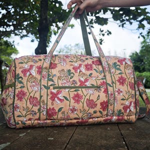 May include: A quilted duffel bag featuring a peach-colored base with a repeating floral pattern in shades of red, pink, and green. The bag includes olive green trim, two top handles, and a horizontal zippered pocket on the front exterior panel.