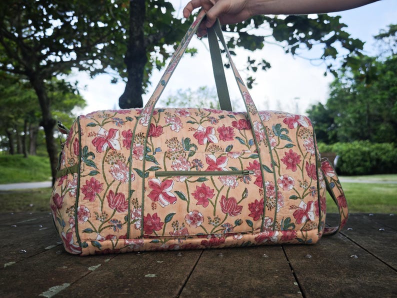 May include: A quilted duffel bag featuring a peach-colored base with a repeating floral pattern in shades of red, pink, and green. The bag includes olive green trim, two top handles, and a horizontal zippered pocket on the front exterior panel.