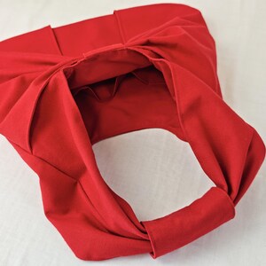 May include: A bright red fabric tote bag with a unique knotted design. The bag features a large opening and a handle made from the same material. The bag is made of a soft, pliable fabric.