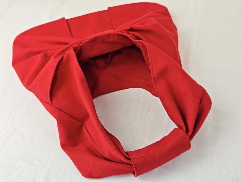 May include: A bright red fabric tote bag with a unique knotted design. The bag features a large opening and a handle made from the same material. The bag is made of a soft, pliable fabric.