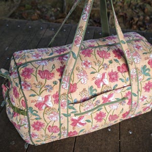 May include: A quilted duffel bag featuring a light peach background with a repeating floral pattern in shades of pink, white, and green. The bag includes two long carrying handles, a zippered top closure, and a small exterior pocket with green trim.