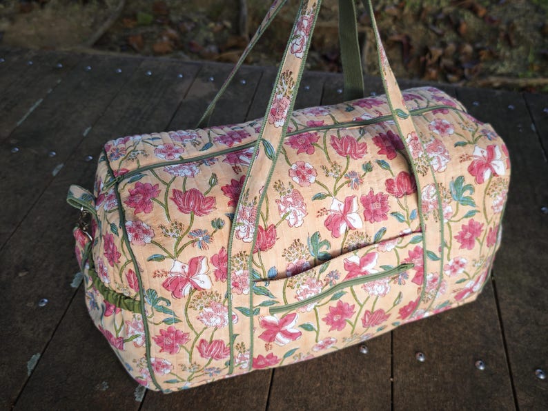 May include: A quilted duffel bag featuring a light peach background with a repeating floral pattern in shades of pink, white, and green. The bag includes two long carrying handles, a zippered top closure, and a small exterior pocket with green trim.