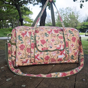 May include: A quilted duffel bag featuring a peach-colored base with a repeating floral pattern in shades of red, pink, and green. The bag includes olive green fabric trim, two top handles, a detachable shoulder strap, and a front flap pocket.