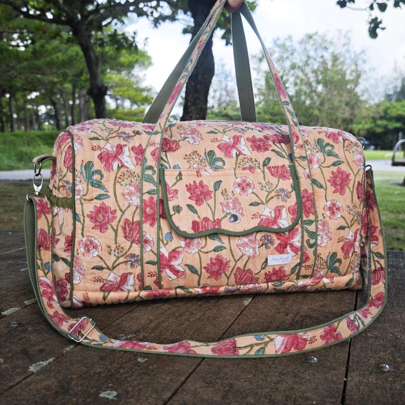 May include: A quilted duffel bag featuring a peach-colored base with a repeating floral pattern in shades of red, pink, and green. The bag includes olive green fabric trim, two top handles, a detachable shoulder strap, and a front flap pocket.
