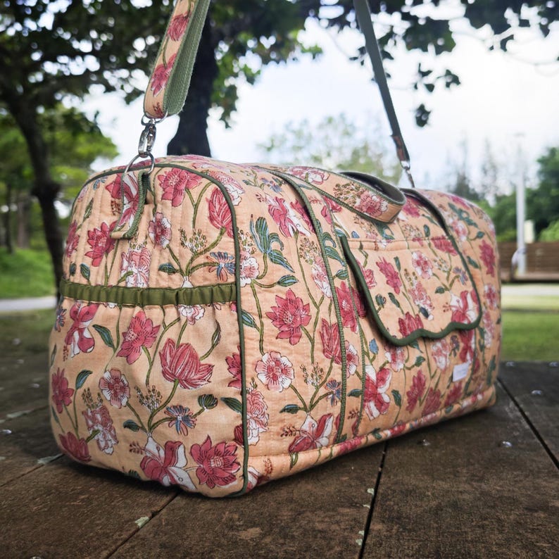 May include: A quilted duffel bag featuring a peach-colored fabric with a repeating floral pattern in shades of pink, red, and green. The bag includes olive green piping, a side pocket with a scalloped flap, and an adjustable shoulder strap.