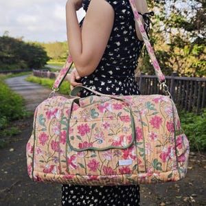 May include: A quilted duffel bag featuring a peach-colored base with a floral pattern in shades of pink, white, and green. The bag includes a rectangular front pocket with a scalloped flap, green piping trim, and a matching adjustable shoulder strap.