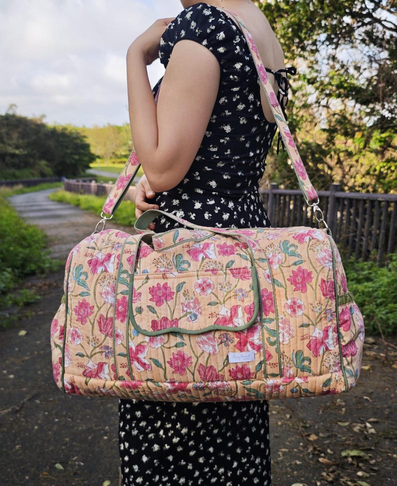 May include: A quilted duffel bag featuring a peach-colored base with a floral pattern in shades of pink, white, and green. The bag includes a rectangular front pocket with a scalloped flap, green piping trim, and a matching adjustable shoulder strap.