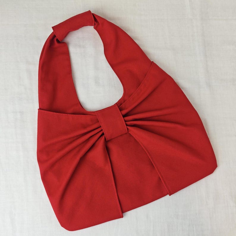 May include: A bright red handbag with a unique bow-like design. The bag features a single handle and a large, gathered bow detail on the front. The bag is made of a smooth, solid-colored fabric.