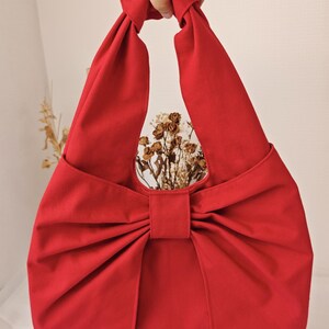 May include: A vibrant red fabric tote bag with a large bow detail. The bag has a knotted handle and a circular opening. The bag is made of a solid red fabric and is empty.