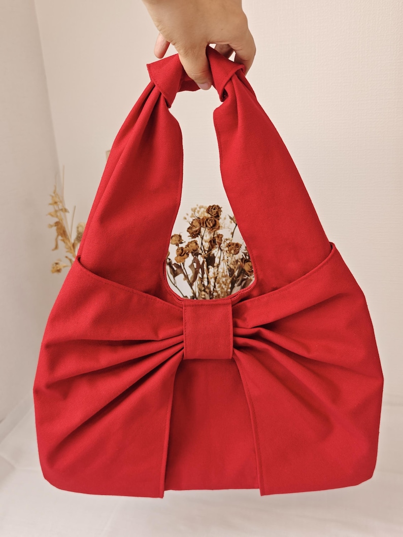 May include: A vibrant red fabric tote bag with a large bow detail. The bag has a knotted handle and a circular opening. The bag is made of a solid red fabric and is empty.