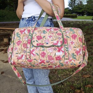 May include: A quilted duffel bag featuring a peach-colored base with a repeating floral pattern in shades of pink, white, and green. The bag includes a front flap pocket with green trim, two top handles, and an adjustable green shoulder strap.