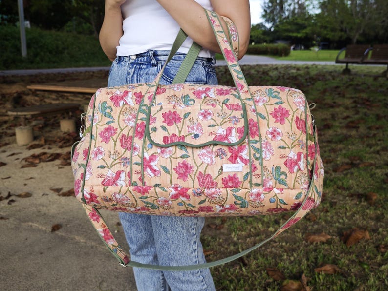 May include: A quilted duffel bag featuring a peach-colored base with a repeating floral pattern in shades of pink, white, and green. The bag includes a front flap pocket with green trim, two top handles, and an adjustable green shoulder strap.
