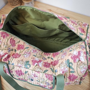 May include: A quilted duffel bag featuring a floral pattern with pink and white flowers on a light orange background. The bag has olive green nylon webbing handles and a matching olive green interior lining with multiple open storage pockets.