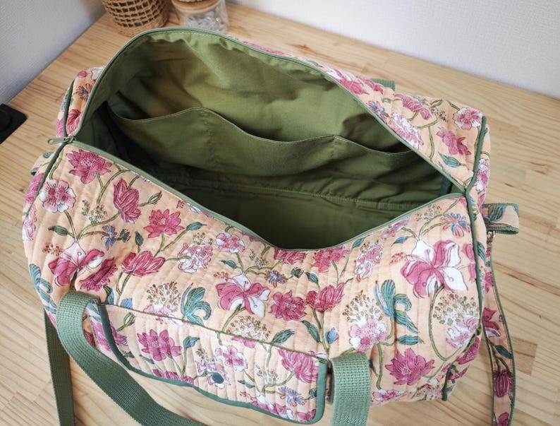 May include: A quilted duffel bag featuring a floral pattern with pink and white flowers on a light orange background. The bag has olive green nylon webbing handles and a matching olive green interior lining with multiple open storage pockets.