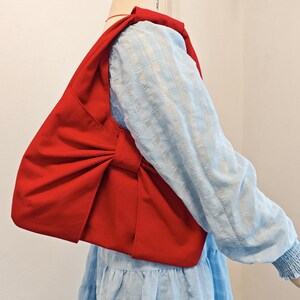 May include: A vibrant red shoulder bag with a large bow detail. The bag is made of a smooth fabric and is worn over a light blue long-sleeved top. The bag's design is simple yet eye-catching.