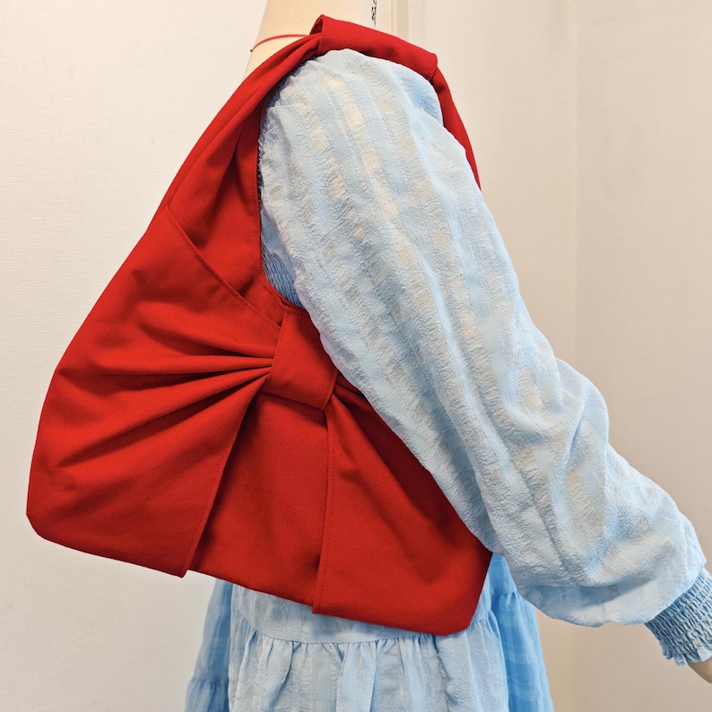 May include: A vibrant red shoulder bag with a large bow detail. The bag is made of a smooth fabric and is worn over a light blue long-sleeved top. The bag's design is simple yet eye-catching.