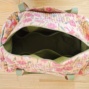 May include: A quilted duffel bag featuring a peach-colored fabric with a pink and green floral pattern. The interior is lined with solid olive green fabric and includes multiple open pockets along the sides. It has olive green webbing handles.