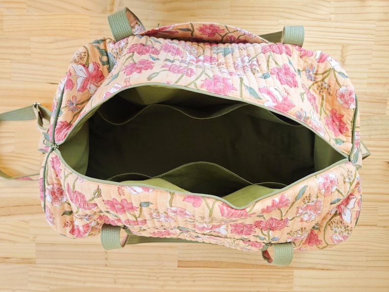 May include: A quilted duffel bag featuring a peach-colored fabric with a pink and green floral pattern. The interior is lined with solid olive green fabric and includes multiple open pockets along the sides. It has olive green webbing handles.