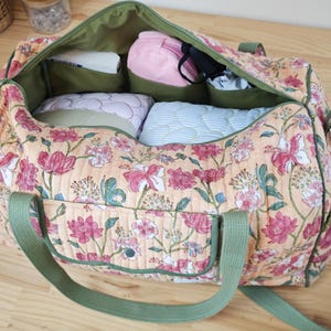 May include: A quilted duffel bag featuring a peach-colored fabric with a pink and green floral pattern. The bag has olive green webbing straps, an exterior pocket with a snap closure, and an open interior lined with olive green fabric.