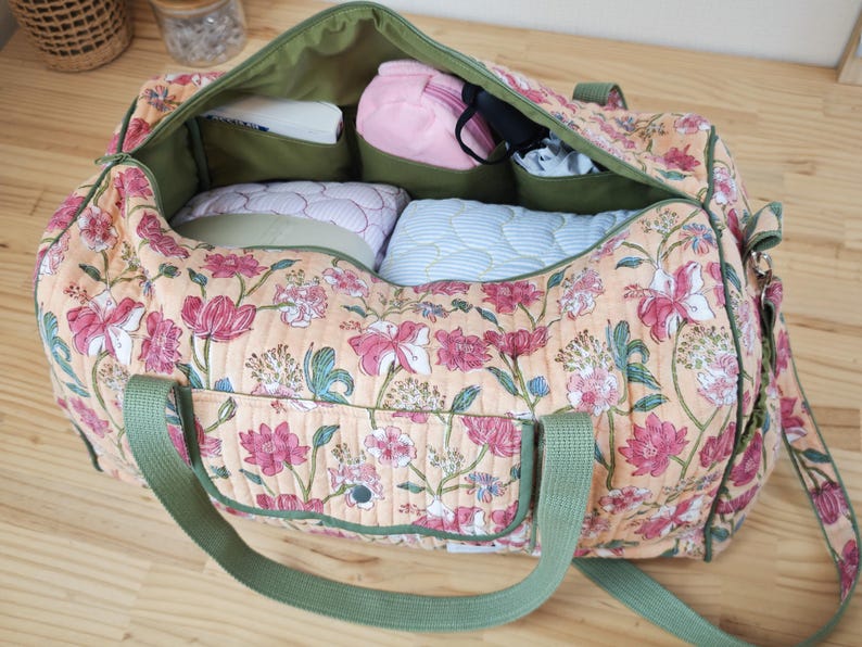 May include: A quilted duffel bag featuring a peach-colored fabric with a pink and green floral pattern. The bag has olive green webbing straps, an exterior pocket with a snap closure, and an open interior lined with olive green fabric.