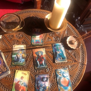 May include: A wooden table with carved details holds a lit candle, incense, and a collection of tarot cards. The cards are spread out, featuring colorful illustrations and titles like "The Lovers," "The Emperor," and "Death."