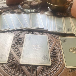 May include: A spread of tarot cards with light green backs and ornate designs, arranged on a carved wooden surface. The cards are laid out in a reading, with several decks visible.