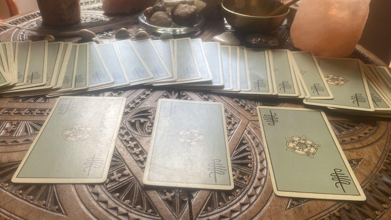 May include: A spread of tarot cards with light green backs and ornate designs, arranged on a carved wooden surface. The cards are laid out in a reading, with several decks visible.