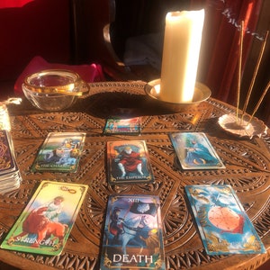 May include: A tarot card reading setup on a carved wooden table. The cards are spread out, featuring images like "The Chariot," "The Emperor," and "Strength." A lit candle, incense, and a glass bowl are also present, creating a mystical atmosphere.