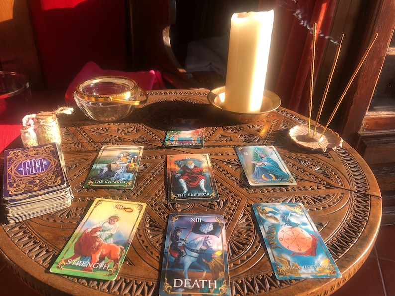 May include: A tarot card reading setup on a carved wooden table. The cards are spread out, featuring images like "The Chariot," "The Emperor," and "Strength." A lit candle, incense, and a glass bowl are also present, creating a mystical atmosphere.