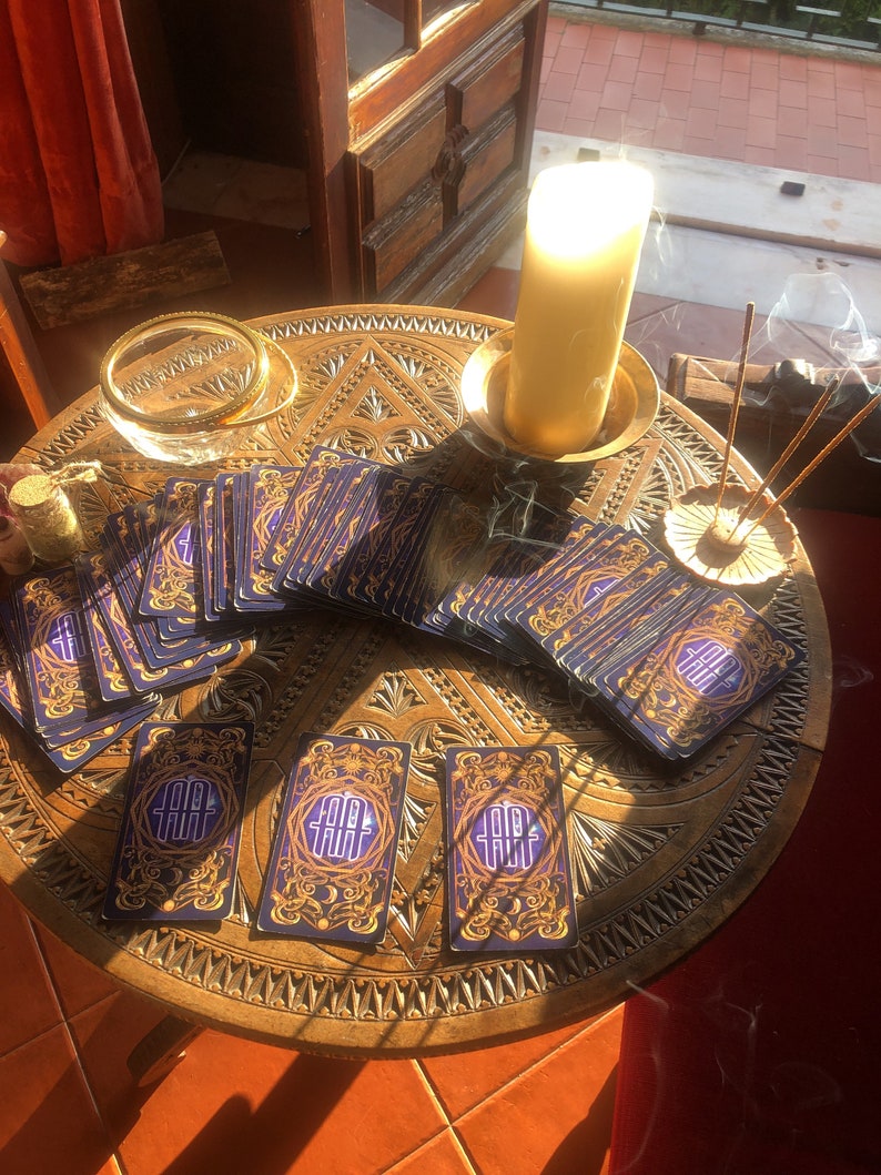 May include: A round, wooden table with intricate carvings holds a deck of tarot cards with blue and gold designs. A lit candle, incense, and a small glass bowl are also on the table. The cards feature the text "TAROT".
