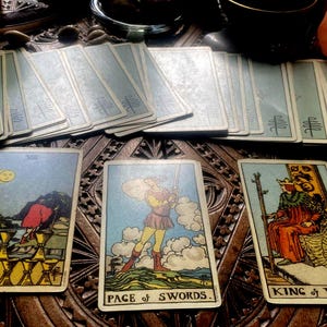 Może przedstawiać: Zestaw kart tarota, z ilustracjami i tytułami takimi jak "Page of Swords" i "King of Wands". Karty ułożone są na rzeźbionej drewnianej powierzchni, z widocznymi kilkoma taliami. Paleta kolorów obejmuje odcienie niebieskiego, żółtego i czerwonego.