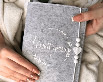 Könnte beinhalten: Grauer Filzbuchumschlag mit einem weißen Kranzdesign und dem Text "Mutterpass" in Weiß. Der Umschlag hat ein weißes Gummiband mit weißen Perlen und einem Herzanhänger.