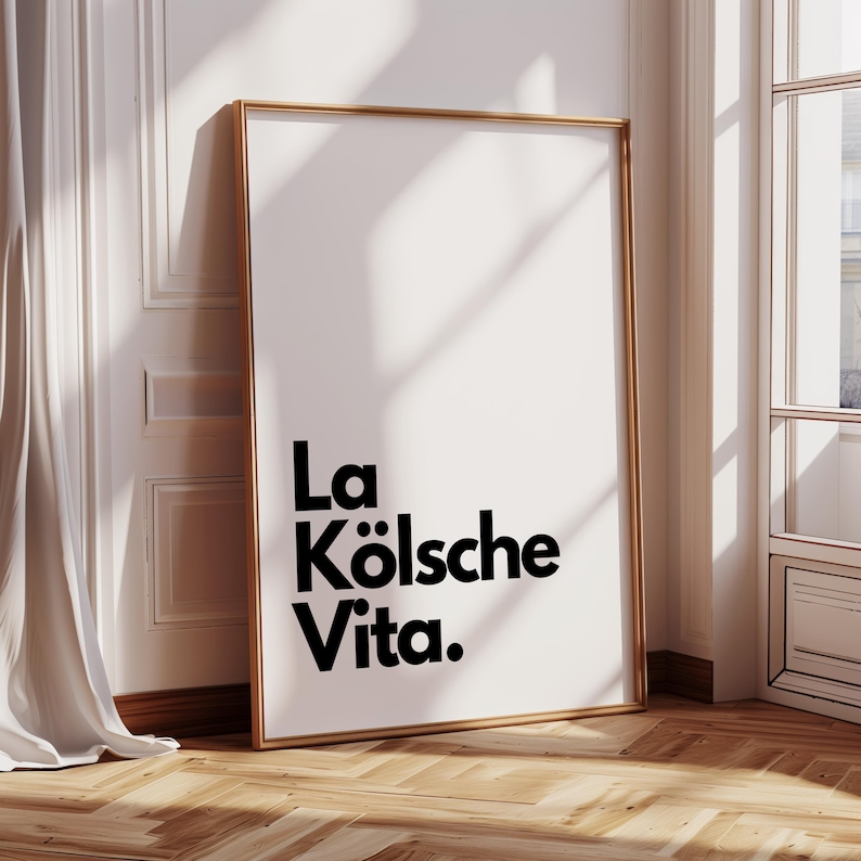May include: A framed art print with the text "La K&ouml;lsche Vita" in black, set against a white background. The frame is gold-colored, and the print leans against a white wall with a wooden floor. A white curtain and window are visible.