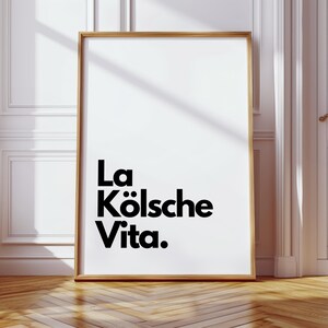 May include: A framed art print with the text "La K&ouml;lsche Vita" in black against a white background. The frame is a light brown color. The print is leaning against a white wall and a wooden floor.