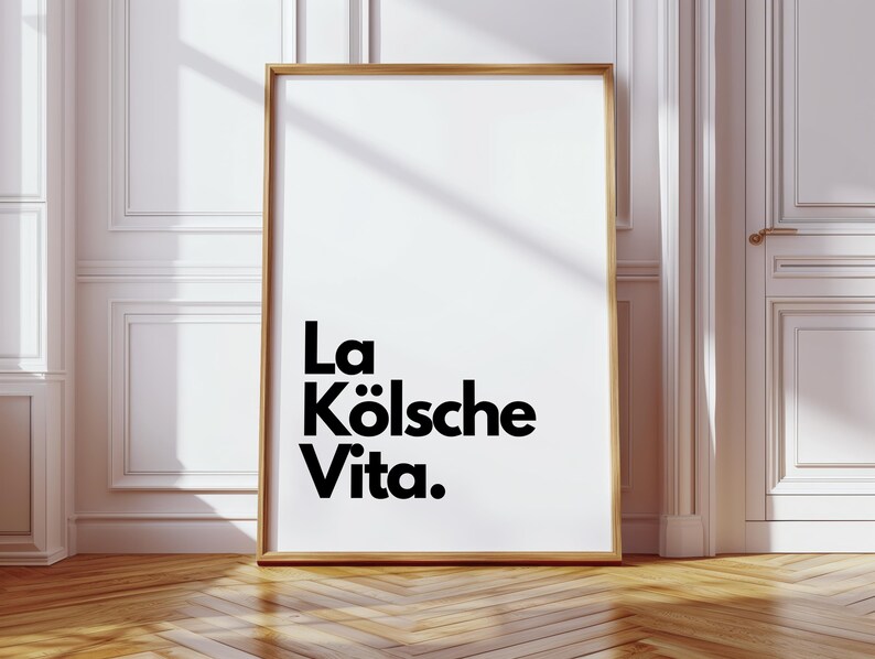 May include: A framed art print with the text "La K&ouml;lsche Vita" in black against a white background. The frame is a light brown color. The print is leaning against a white wall and a wooden floor.
