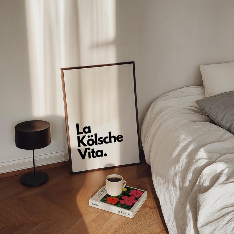 May include: A framed art print with the text "La K&ouml;lsche Vita" in black. A small black lamp, a coffee cup, and a book with colorful flowers are on the floor. A bed with white linens is in the background.
