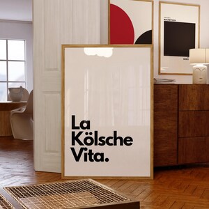 May include: A framed art print with the text "La K&ouml;lsche Vita" in black. The print is in a room with wooden floors and a wooden table. Two other framed art prints are visible in the background.