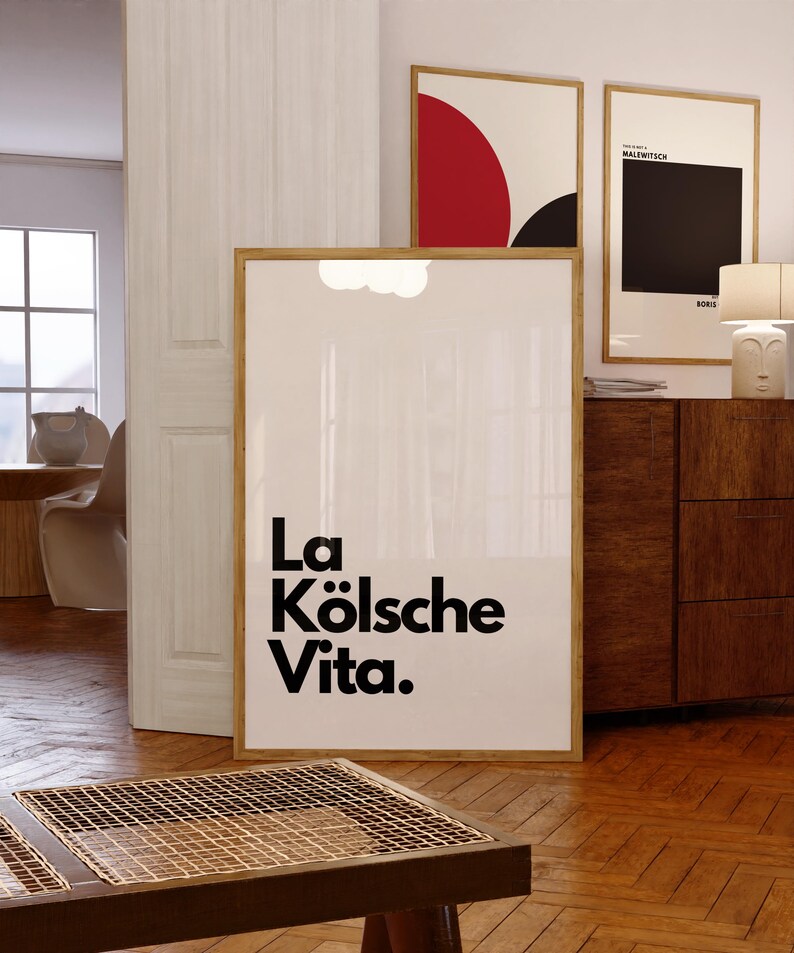May include: A framed art print with the text "La K&ouml;lsche Vita" in black. The print is in a room with wooden floors and a wooden table. Two other framed art prints are visible in the background.