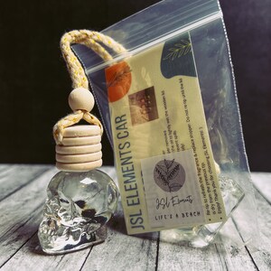 K&ouml;nnte beinhalten: Ein klarer, sch&auml;delformiger Auto-Lufterfrischer mit einem Holzdeckel und einem gelb-wei&szlig;en Seil. Das Produkt ist in einer transparenten Plastikt&uuml;te mit der Aufschrift "JSL ELEMENTS CAR" und "LIFE'S A BEACH" verpackt.