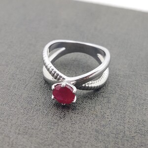 Op de afbeelding: Een zilveren ring met een kruisontwerp. De ring heeft een ronde, helderrode edelsteen in het midden. De band heeft aan één kant een textuurpatroon. De ring is geplaatst tegen een grijze achtergrond.