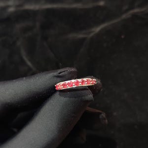 Peut inclure: Une bague en argent ornée d'une rangée de petites pierres précieuses rondes et rouges. La bague est présentée sur un fond sombre, mettant en valeur la couleur rouge vif des pierres. La bague semble être de style alliance.