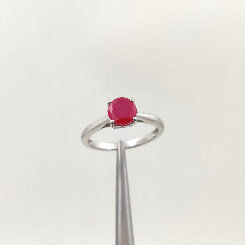Puede incluir: Un anillo de plata con una gema roja redonda y vibrante. El anillo tiene una banda simple y un engaste de cuatro puntas que sujeta la gema de forma segura. El anillo es sostenido por una herramienta plateada sobre un fondo blanco.