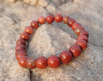 May include: A red jasper beaded bracelet. The bracelet is made of round, polished beads in shades of red and brown. The bracelet is resting on a gray rock surface.