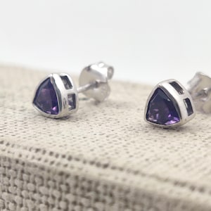 May include: A pair of stud earrings with a triangular, deep purple gemstone set in a silver-colored metal frame. The earrings are displayed on a textured, light-colored surface, showcasing their design and color.